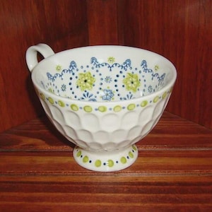 Peut inclure: Tasse &agrave; th&eacute; en c&eacute;ramique blanche avec une anse, orn&eacute;e de motifs floraux bleus et jaunes. La tasse a un corps arrondi textur&eacute; et un petit socle, &eacute;galement d&eacute;cor&eacute; d'accents bleus et jaunes. La tasse est pos&eacute;e sur une surface en bois.