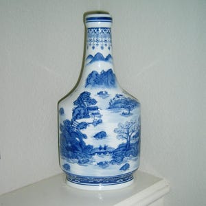 May include: A blue and white porcelain vase with a landscape design. The vase features a bottle shape with a wide base and a narrow neck. The landscape design includes trees, mountains, and bridges. The vase is approximately 12 inches tall.