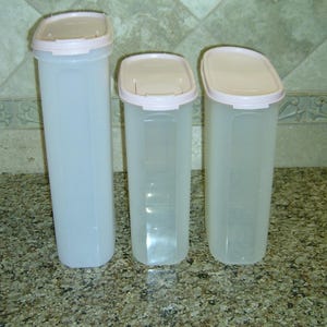May include: Three tall, rectangular food storage containers made of translucent plastic with rounded corners. Each container features a light pink, snap-on lid with a flip-top opening mechanism. The containers vary in height, with one taller unit.