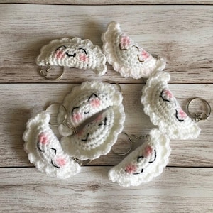 May include: Several white crochet dumpling keyrings with smiling faces, featuring pink blush and black stitched details. The keyrings are arranged on a light wooden surface.