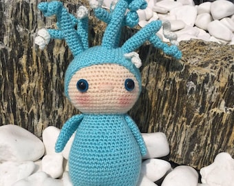May include: A handmade crochet doll in a whimsical design. The doll features a light blue body and head covering, with blue and white accents on the head. It has large blue eyes and a light pink blush. The doll is sitting on white stones.