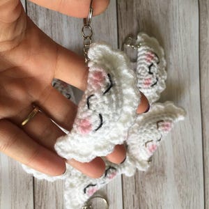 Puede incluir: Llaveros de ganchillo blancos hechos a mano con forma de empanadillas sonrientes, con mejillas rosas y ojos cosidos negros. Cada llavero tiene un llavero plateado adjunto. Varios llaveros se sostienen en una mano, con un fondo de madera.