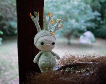 May include: A handmade crochet doll in the shape of a whimsical tree-like creature. The doll is primarily light green with a rounded body, small arms, and a head with multiple upward-reaching branches adorned with orange and yellow accents. It has large black eyes and a sweet expression.