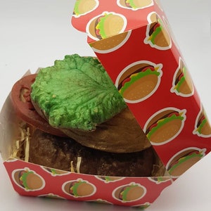 Puede incluir: Un artículo novedoso con forma de hamburguesa, con una hamburguesa marrón, una capa de lechuga verde y una rodaja de tomate roja, presentado en una caja roja con temática de hamburguesa. La caja tiene un patrón de hamburguesas repetido.