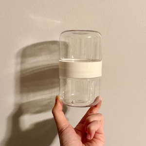 May include: A clear, cylindrical glass container featuring a smooth, opaque white band encircling the center. The vessel consists of two distinct, stackable glass sections joined by this central white ring, creating a sleek, minimalist storage unit.