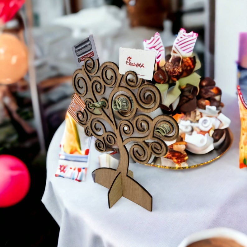 May include: A wooden money tree with a swirling design, holding cash and gift cards from Chipotle and Chick-fil-A. The tree is on a white tablecloth, with a blurred background of food and decorations.