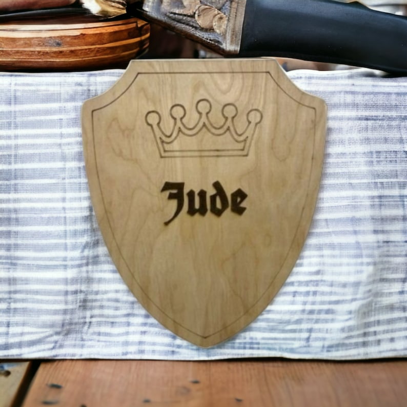 May include: A wooden shield with a crown and the name "Jude" engraved on it. The shield is light brown and has a dark brown border.