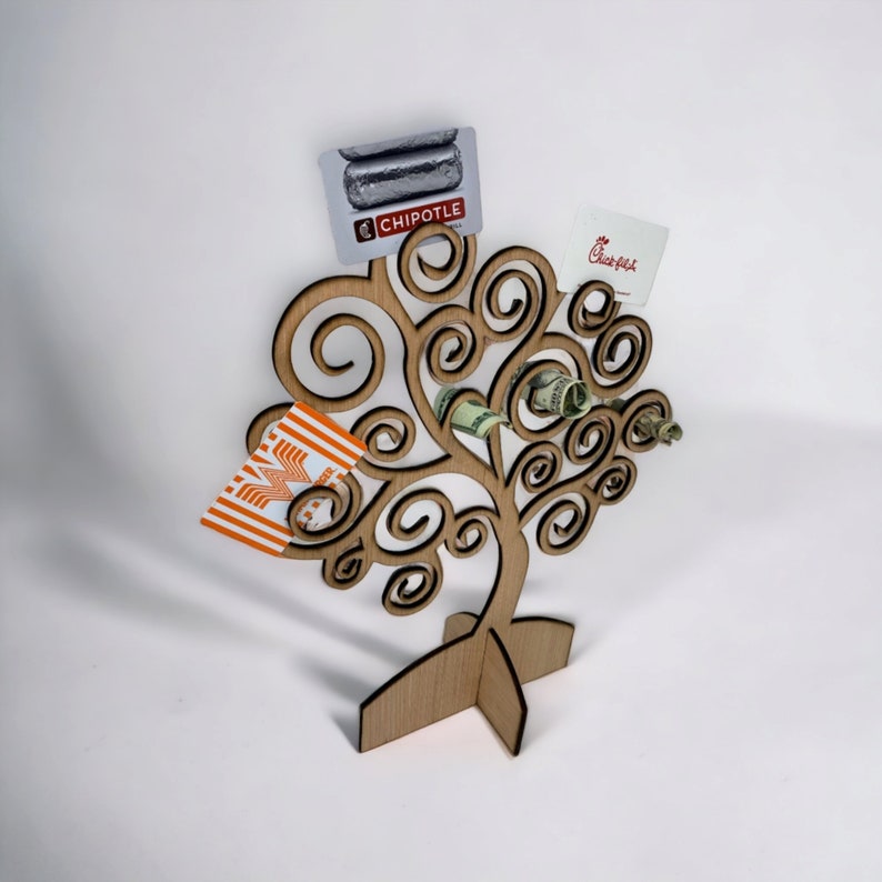 May include: A wooden tree-shaped gift card and money holder. The holder has a swirling branch design and holds gift cards from Chipotle, Chick-fil-A, and Whataburger, along with rolled-up dollar bills. The holder is made of light-colored wood.