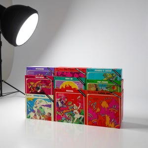 May include: A stack of vintage vinyl record albums with colorful, psychedelic cover art. The album covers feature illustrations and text, including titles like "Festival" and "Flaming Pops." The records are stacked on a white surface.
