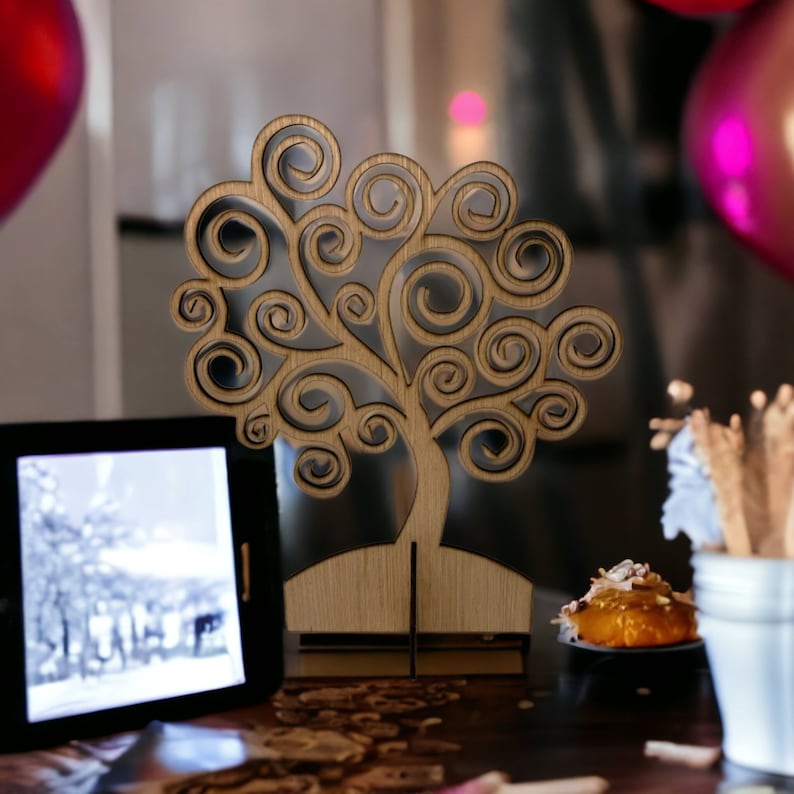 May include: A wooden tree-shaped decorative item with swirling branches. The tree is light brown and stands on a small base. The background includes a tablet displaying a black and white image, a small dessert, and balloons.