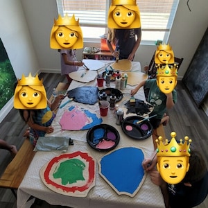 May include: Children paint wooden shields with different colors of paint. The shields are shaped like a rounded rectangle and have a raised edge. The children are wearing crowns.