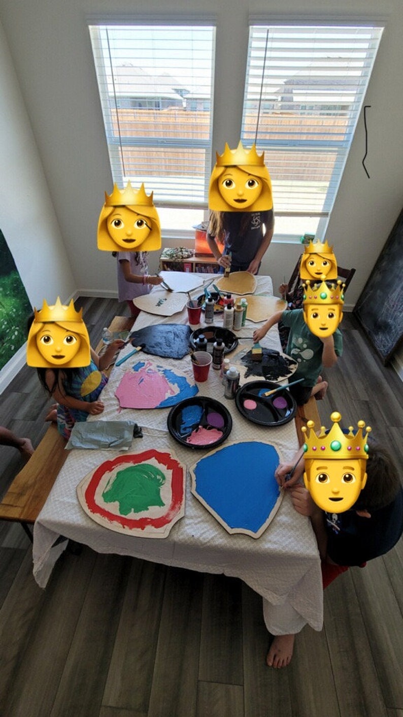 May include: Children paint wooden shields with different colors of paint. The shields are shaped like a rounded rectangle and have a raised edge. The children are wearing crowns.