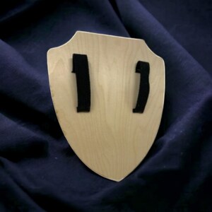 May include: A wooden shield with a light brown finish and two black straps on the back.