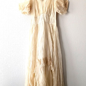 May include: A cream-colored floor-length dress featuring short puff sleeves, a V-neckline, and intricate lace detailing along the bodice and shoulders. The skirt is constructed from layered, sheer, crinkled fabric with lace trim accents throughout.
