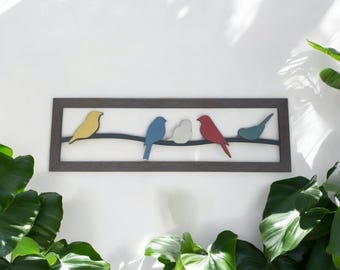 May include: A rectangular wall art piece featuring a dark brown frame enclosing a white background. Five stylized birds in yellow, blue, white, red, and teal are perched on a thin, dark horizontal line that spans the width of the frame.
