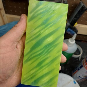 May include: A rectangular block of translucent material with a vibrant green and yellow color scheme. The surface features a wavy pattern of darker green streaks, creating a dynamic visual effect. The block is held in a hand, showcasing its size and texture.