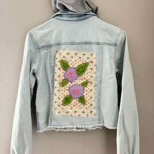 May include: Light blue denim jacket with a gray hood. The back features a floral patch with two purple flower appliqués, green leaves, and a floral patterned background. The jacket has long sleeves and a frayed hem.