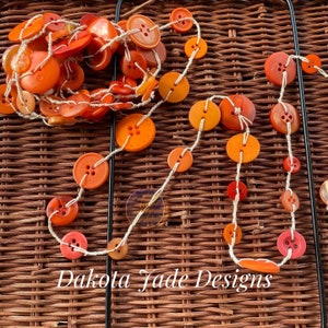 May include: A string of orange and red buttons strung on white twine. The buttons are various sizes and shapes. The string is hanging on a woven wicker surface.