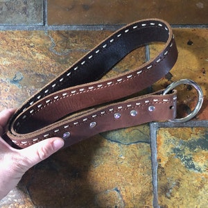 May include: A brown leather belt with white stitching and silver studs. The belt has a silver ring buckle.