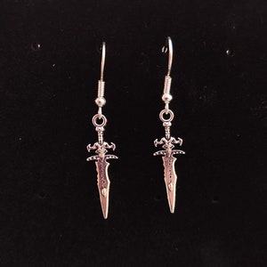 May include: A pair of silver sword-shaped earrings with a detailed design. The earrings are hanging from silver hooks.