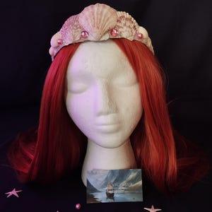Peut inclure: Une couronne de coquillages avec des perles roses et une perruque rousse sur une tête de mannequin blanche. La couronne est faite de divers coquillages et ornée de petites perles roses. La perruque a de longs cheveux roux. Le fond est sombre.