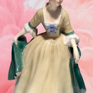 Peut inclure: Une figurine en porcelaine d'une femme en robe jaune avec un châle vert. Elle porte un ruban bleu dans les cheveux et a une expression faciale délicate.