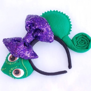 May include: Purple sequined bow with a green felt chameleon character on a black headband. The chameleon has a curled tail and large eyes.