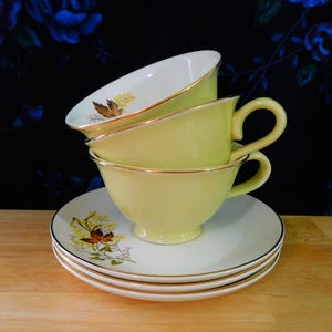 May include: A stack of vintage teacups and saucers. The cups are a pale yellow with gold trim and handles. The saucers are white with gold trim and a floral design. The background is a dark blue floral pattern.