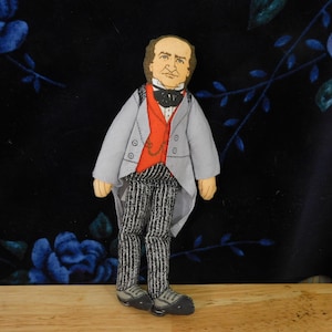 May include: A handmade doll of a man in a gray coat, red vest, and striped pants. The doll has a black bow tie and a pocket watch. The background is a dark blue floral pattern.