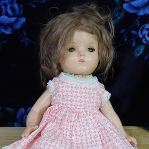 May include: Vintage doll with brown hair and a pink floral dress. The doll has a white lace collar and is set against a dark blue floral background. The doll's face is painted with detailed features.
