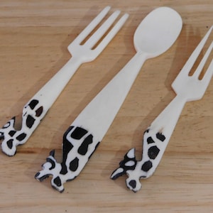 May include: A set of three white utensils: two forks and a spoon. The handles feature a black and white spotted design. The utensils are arranged on a light brown wooden surface.