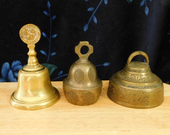 May include: Three antique brass bells of varying shapes and sizes. The bell on the left has a handle with a circular emblem. The middle bell is round with leaf designs. The bell on the right has a handle and etched patterns.
