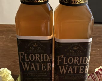 May include: Two tall, clear glass bottles filled with amber-colored Florida Water spiritual cologne. Each bottle features a black cap with a gold band and a black label with white text reading Ms. G's Curious Goods Florida Water Spiritual Cologne.
