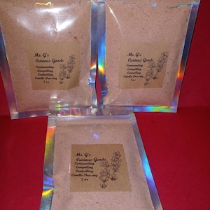 Pode incluir: Três pequenos sacos de plástico transparente cheios de um pó castanho claro. Cada saco tem um rótulo castanho com o texto "Ms. G's Curious Goods Commanding Compelling Controlling Candle Dressing 2 oz".