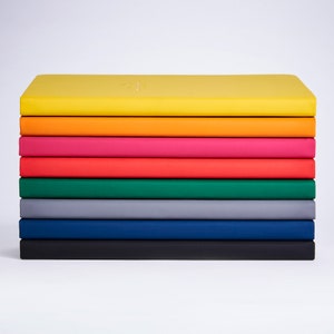 Może przedstawiać: Stosik dziesięciu zeszytów w różnych kolorach, w tym żółtym, pomarańczowym, różowym, czerwonym, zielonym, szarym i niebieskim. Zeszyty są wszystkie zamknięte i ułożone jeden na drugim.