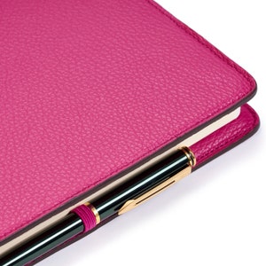 Può includere: Un diario rilegato in pelle rosa con una penna dorata infilata nella fascia elastica.