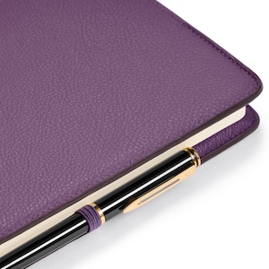 Puede incluir: Un cuaderno de cuero morado con un bolígrafo negro metido en la portada.