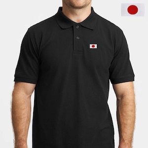Op de afbeelding: Een zwart poloshirt met korte mouwen, een klassieke kraag, een knoopsluiting en een kleine rechthoekige patch op de linkerborst met de nationale vlag van Japan, bestaande uit een rode cirkel op een witte achtergrond.