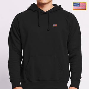 Op de afbeelding: Een zwarte hoodie met lange mouwen en een kleine geborduurde Amerikaanse vlag op de linkerborst. De trui is voorzien van een kangoeroezak, een capuchon met trekkoord en geribbelde boorden aan de mouwen en de onderkant van het kledingstuk.