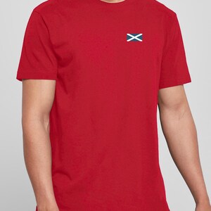 Op de afbeelding: Een rood T-shirt met een klein geborduurd Schots vlaggetje op de linkerborst. De vlag toont een witte X-vormige kruis op een blauwe achtergrond. Het shirt heeft korte mouwen en is gemaakt van effen rode stof.