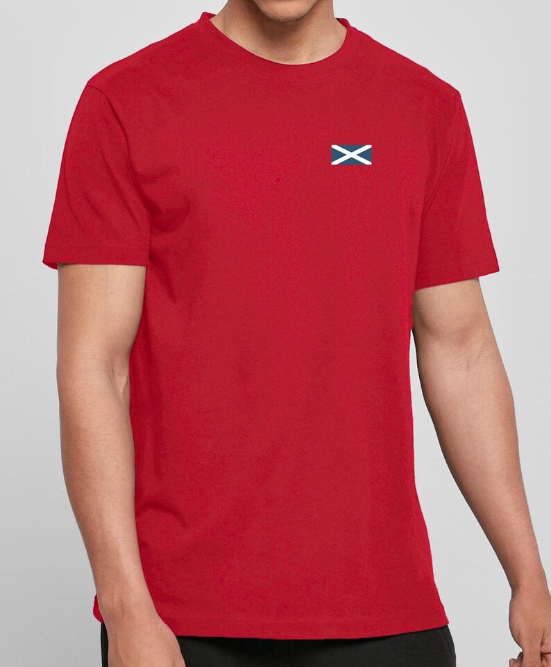 Op de afbeelding: Een rood T-shirt met een klein geborduurd Schots vlaggetje op de linkerborst. De vlag toont een witte X-vormige kruis op een blauwe achtergrond. Het shirt heeft korte mouwen en is gemaakt van effen rode stof.