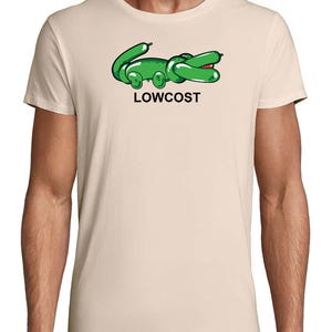 May include: Beige t-shirt featuring a green cartoon alligator design with the word "LOWCOST" in black text below. The alligator has a balloon-like appearance with red accents on its mouth.