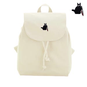 Op de afbeelding: Een crèmekleurige canvas rugzak met een geborduurde patch van een zwarte kat die een mes vasthoudt met rode bloeddetails. De tas heeft een bovenflap, een trekkoordsluiting met twee geknoopte koorden en een handvat aan de bovenzijde.