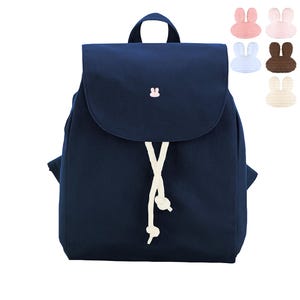 Könnte beinhalten: Ein marineblauer Rucksack mit Klappverschluss und einem kleinen gestickten rosa Hasensymbol. Die Tasche verfügt über eine weiße Kordel mit verknoteten Enden. Fünf kleine, separate hasenförmige Aufnäher in Rosa, Weiß, Braun und Creme sind abgebildet.