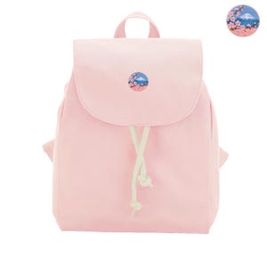 Op de afbeelding: Een lichtroze rugzak met een ronde patch op de voorflap. De patch toont een blauwe bergtop omringd door roze kersenbloesems. De tas heeft een handvat aan de bovenkant, een overslagflap en witte trekkoorden met geknoopte uiteinden.