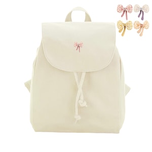 Könnte beinhalten: Ein cremefarbener Rucksack mit einer oberen Klappe, die mit einer kleinen gestickten rosa Schleife verziert ist. Das Hauptfach wird mit einem Kordelzug geschlossen. Vier weitere Schleifenoptionen in Rosa, Blau, Gelb und Pfirsich sind abgebildet.