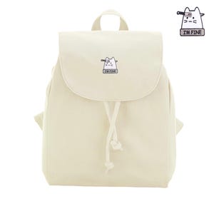 May include: A cream-colored canvas backpack featuring a small embroidered patch of a white cat with a knife stuck in its head and the text I'M FINE. The bag has a fold-over flap, a drawstring closure with knotted ends, and a top carry handle.