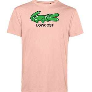 May include: A light pink t-shirt featuring a green cartoon alligator design with the word "LOWCOST" in black text. The alligator has a red mouth and is in a relaxed pose. The shirt has short sleeves.