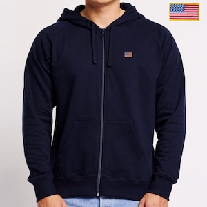Op de afbeelding: Een marineblauwe hoodie met rits en een kleine geborduurde Amerikaanse vlag op de linkerborst. Het kledingstuk heeft een capuchon met trekkoord, lange mouwen met geribbelde boorden en twee voorzakken. Een grotere vlagpatch is ook afgebeeld.