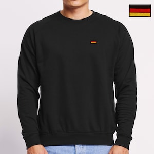 May include: A black long-sleeve sweatshirt featuring a small embroidered German flag on the left chest. The garment has a crew neckline, ribbed cuffs, and a ribbed hem. The flag consists of three horizontal stripes in black, red, and gold.